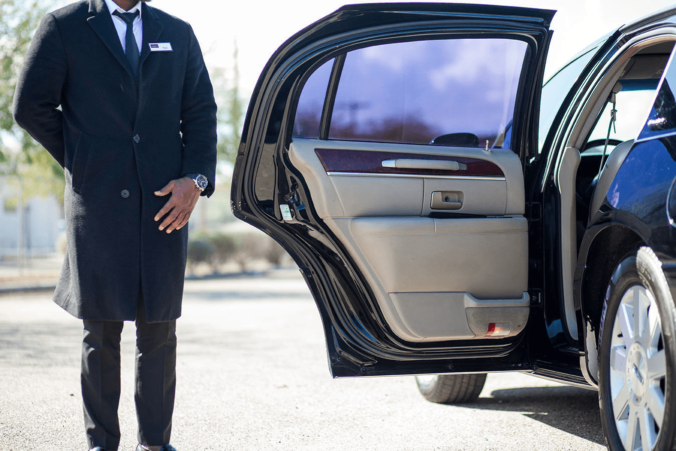 Professional chauffeur in dark suit standing beside black luxury sedan with rear passenger door open, showing premium beige leather interior